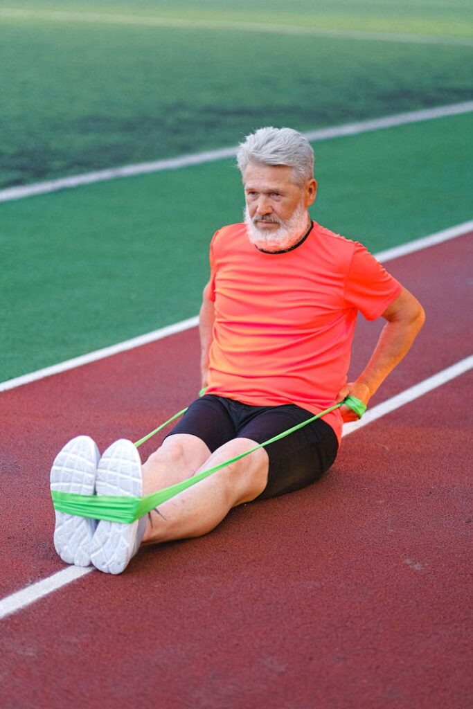 Full body of elderly male stretching and warming up with elastic rubber on racetrack in stadium